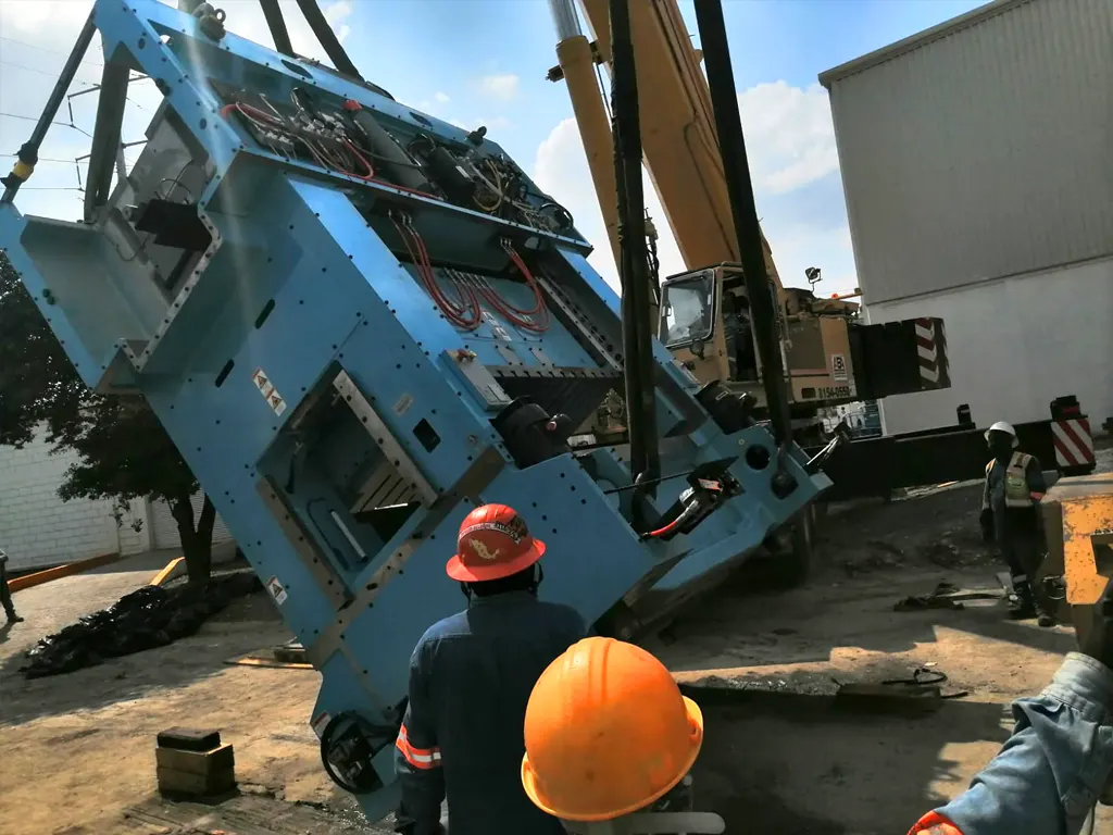 Trabajadores con casco supervisando la manipulación de maquinaria industrial con grúa